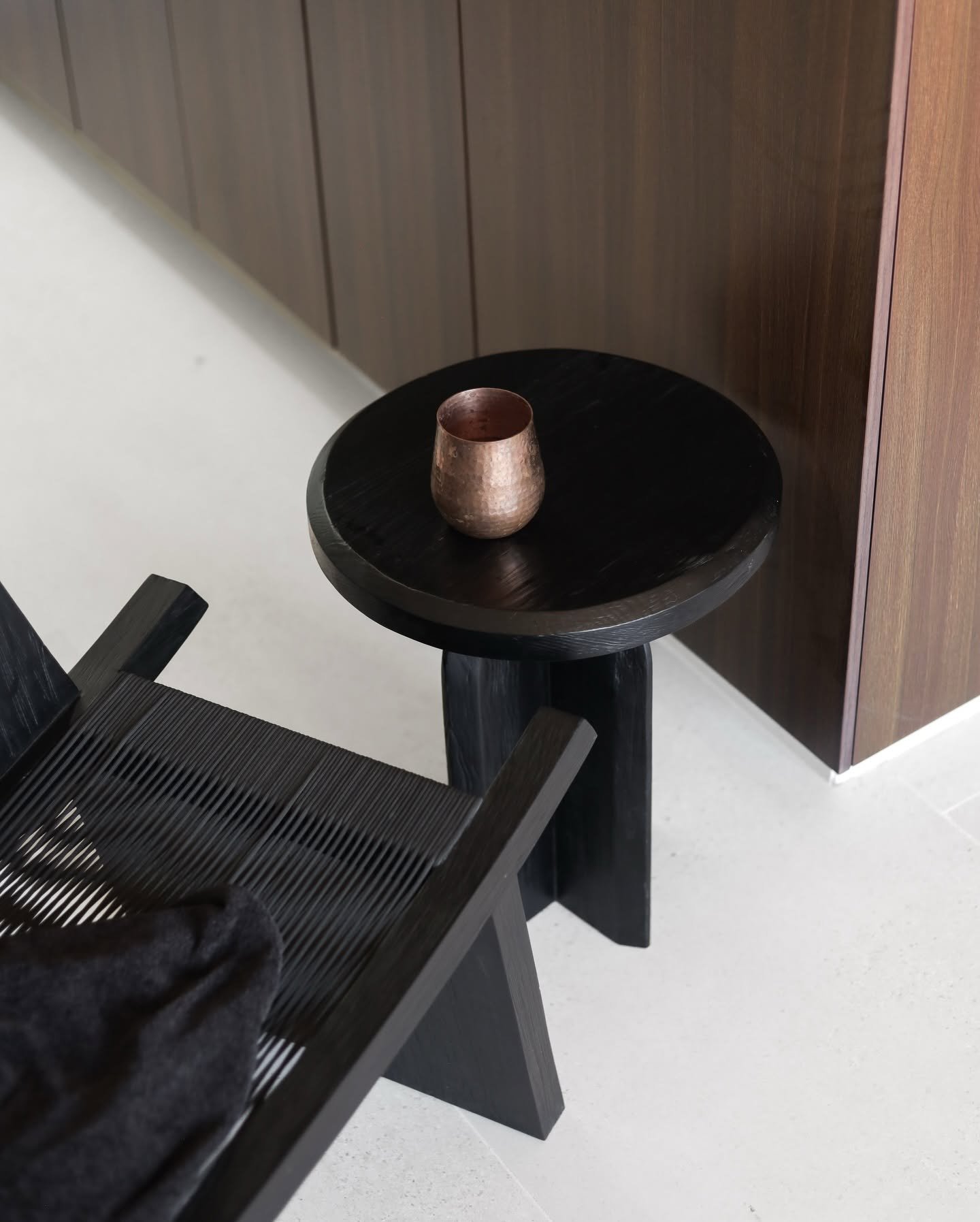 Black carved side table with a hammered copper cup beside a woven lounge chair